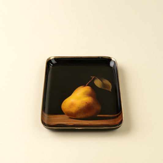 A square black ceramic plate with a glossy finish, featuring a printed still life of a yellow pear with a leaf on a wooden surface. The plate is photographed on a clean, solid, warm off-white background.