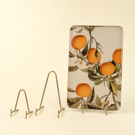 A rectangular decorative enamel tray with a vintage-style print of oranges on branches, displayed on a small metal stand against a warm cream background.