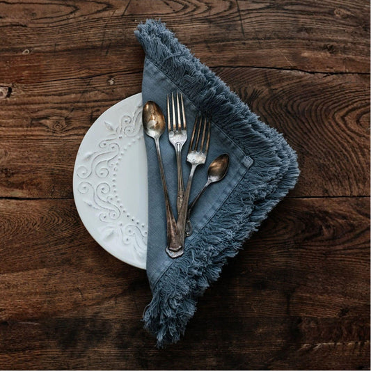 A folded fringed napkin in a muted blue-gray color, styled on top of a white decorative plate with silver cutlery, set on a rustic dark wood table.