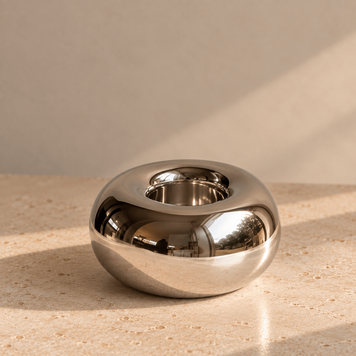 A polished chrome orb-shaped tealight holder, set on a beige stone surface against a matching beige wall with soft diagonal shadows.