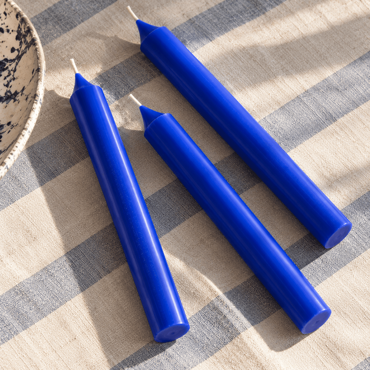 Three cobalt blue taper candles arranged diagonally on a beige and blue striped fabric surface.