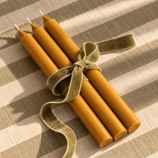 Four yellow ochre taper candles tied together with a wide olive-green velvet ribbon, arranged on a beige and olive striped fabric background.