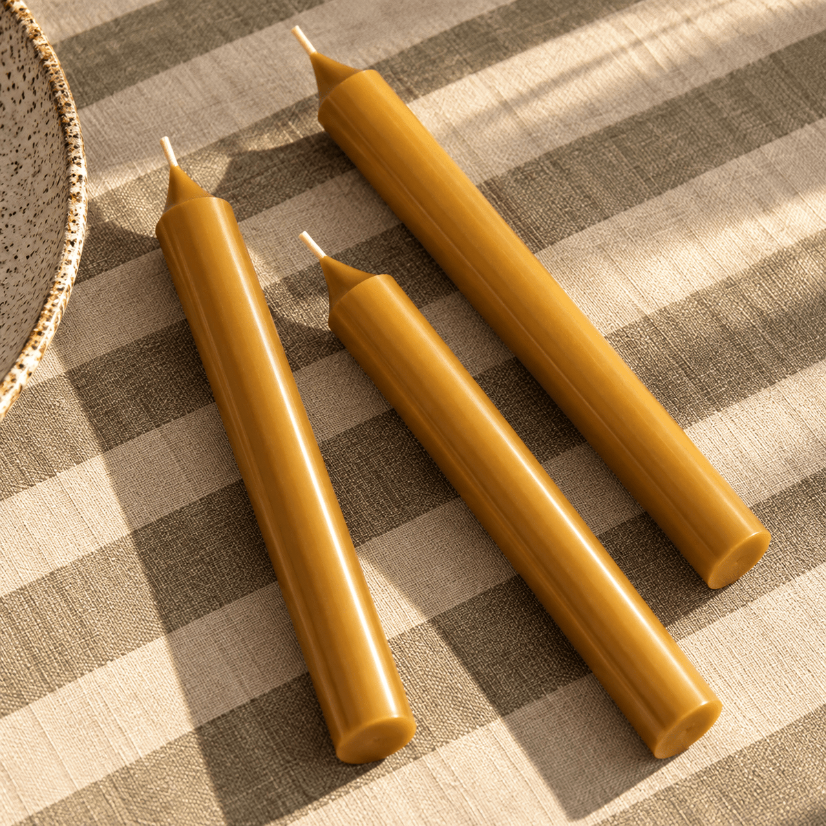 Three tapered yellow ochre candles arranged diagonally on a beige and grey checkered fabric surface with warm sunlight and shadows.