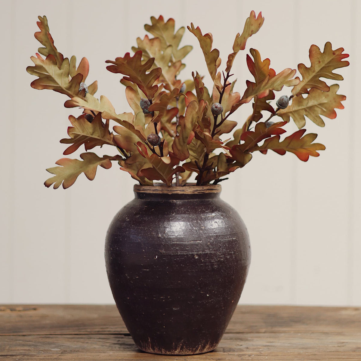 Artificial oak leaf arrangement in a dark brown ceramic vase, displayed on a rustic wooden surface against a light beige paneled wall.