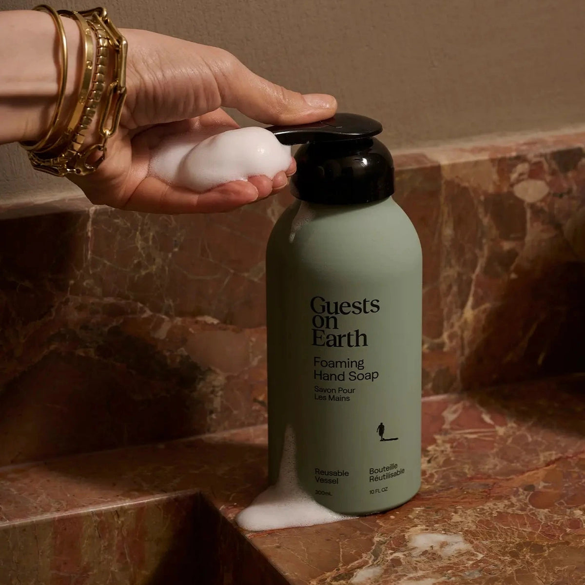A bottle of Guests on Earth Foaming Hand Soap sits on a brown marble countertop with a hand dispensing white foam into a sink. The bottle is a muted sage green with a black pump.