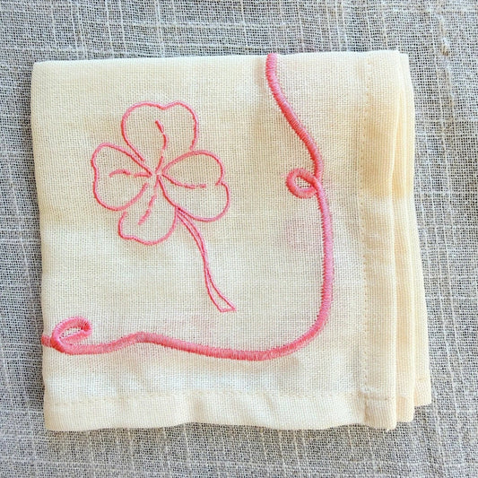 A folded cream-colored linen napkin featuring a pink embroidered four-leaf clover and a matching pink border design, displayed on a textured gray linen background.