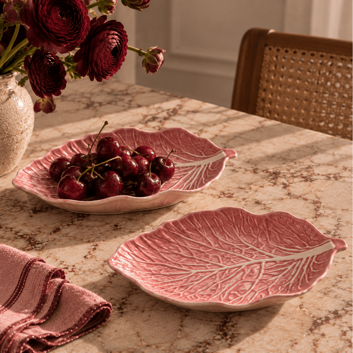 Two pink cabbage leaf ceramic serving platters on a marble table, one filled with cherries and the other empty, with a floral arrangement and woven chair in the background.