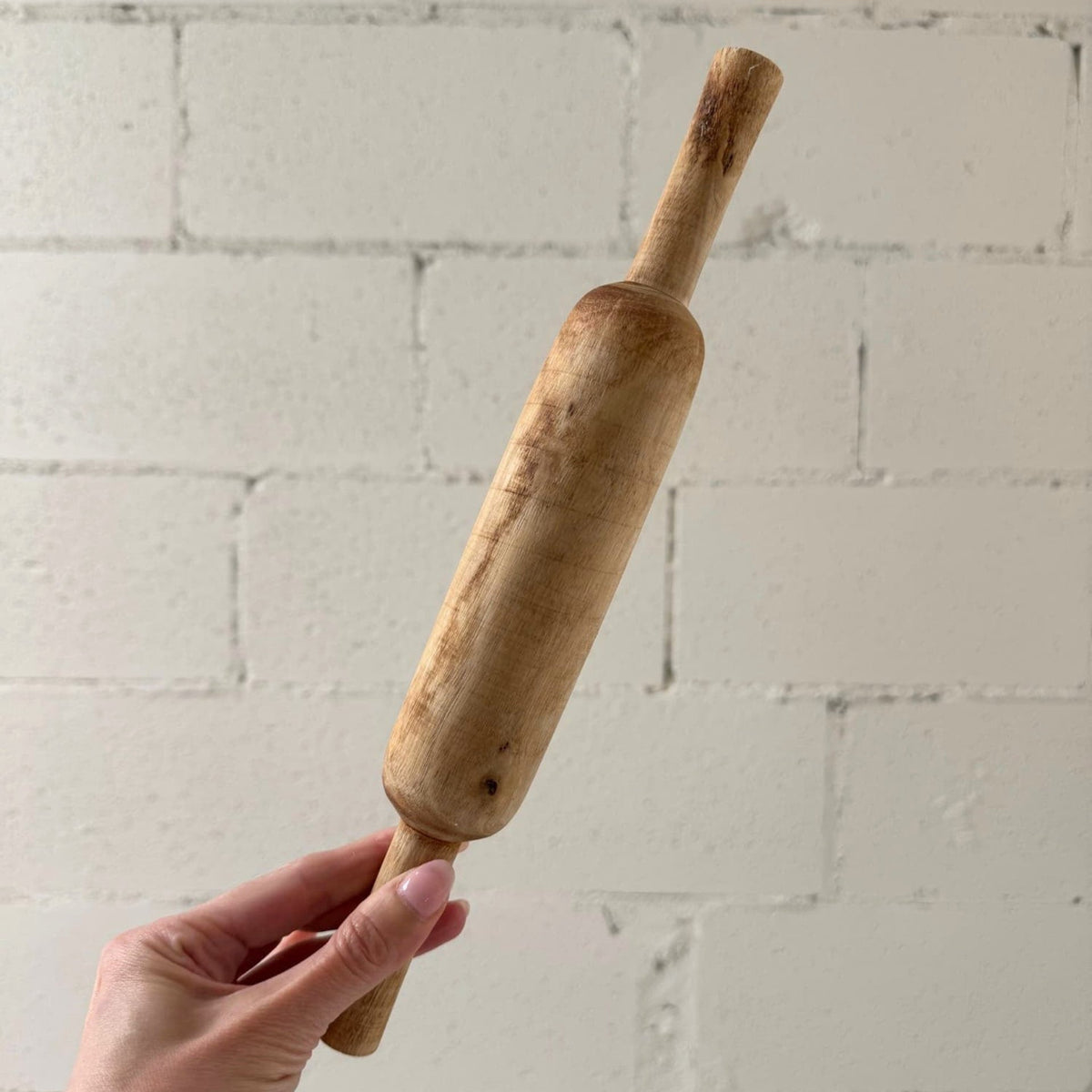 A hand holding a small wooden rolling pin against a white painted brick wall background.