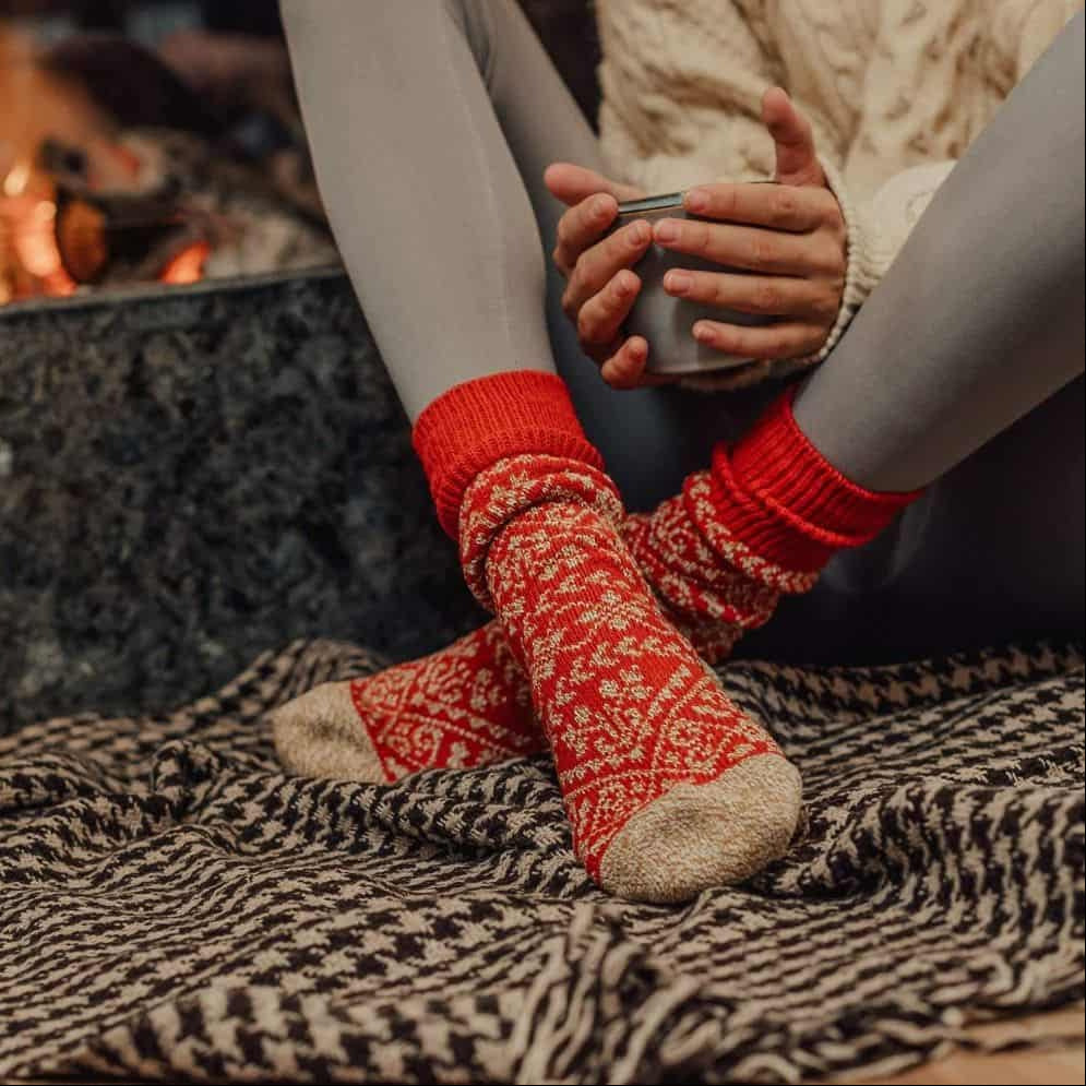 Person wearing red socks with a pattern, sitting by a fireplace holding a mug.