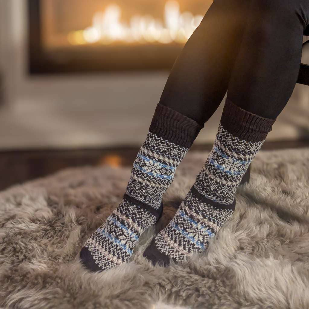 A person seated by a fireplace wearing dark leggings and patterned knit socks with a Fair Isle snowflake design in gray, white, and light blue, sitting on a shag rug.