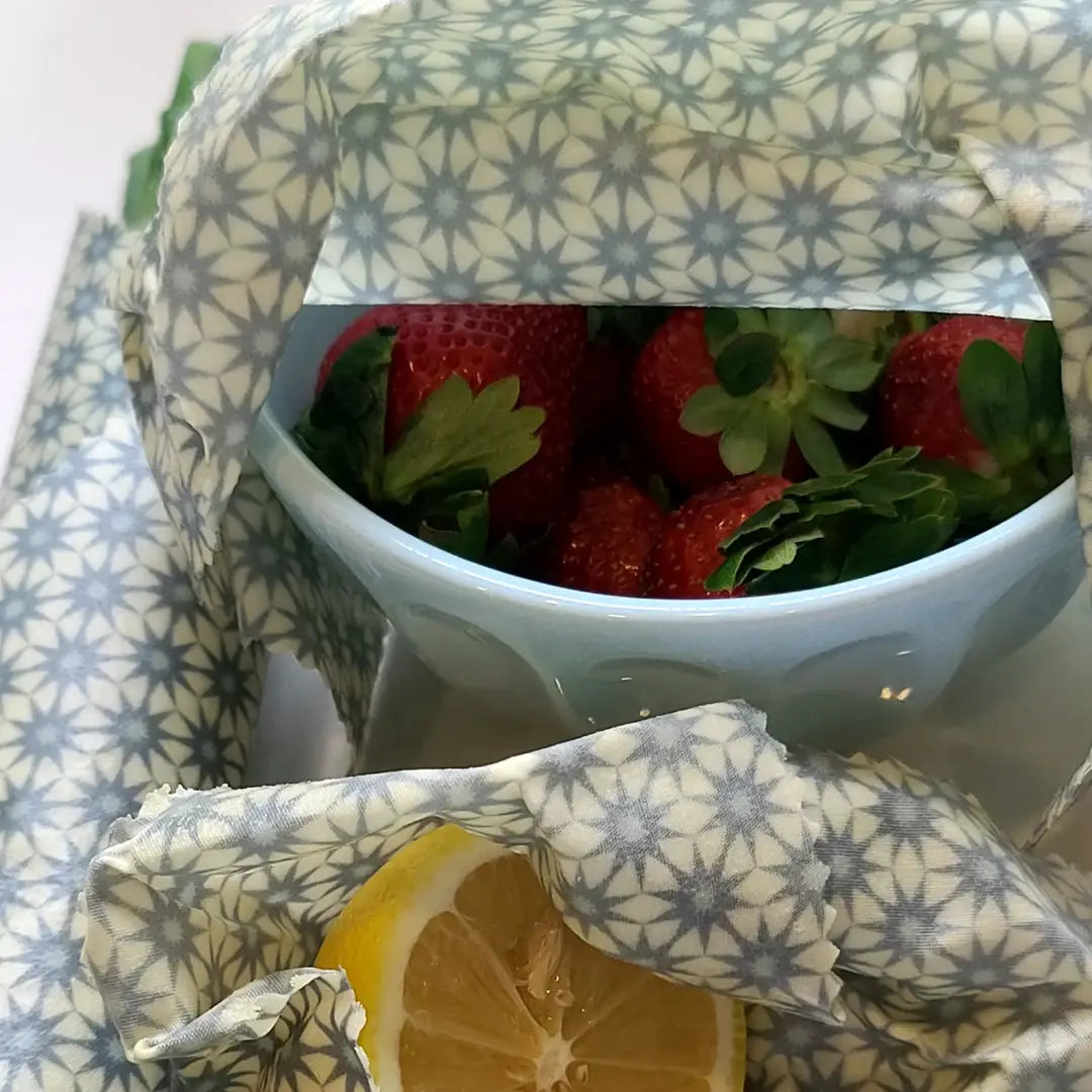 A close-up of reusable patterned fabric beeswax cloth wrap, draped around a bowl of fresh strawberries and a lemon slice. The fabric features a repeating gray and blue starburst pattern on a warm cream background.