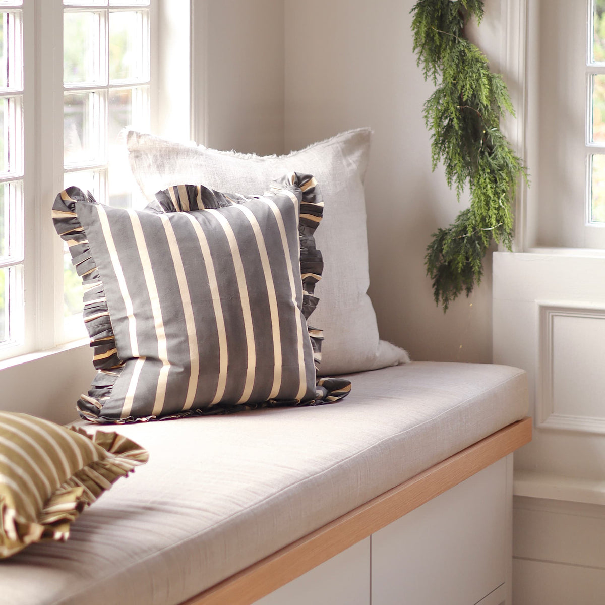 A cozy window seat with neutral-toned cushions, including a striped gray and cream pillow and a white pillow, styled with a green garland on a white wall in a bright room.