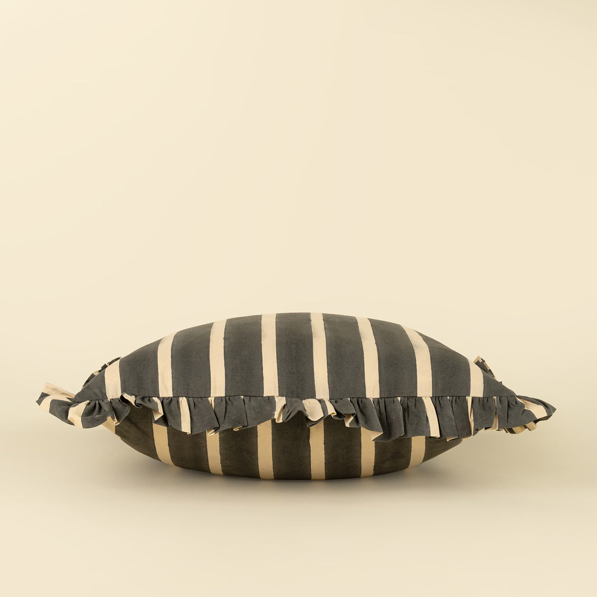 A striped throw pillow with dark gray and cream vertical stripes, featuring a ruffled fabric trim on the sides, set against a solid cream background.