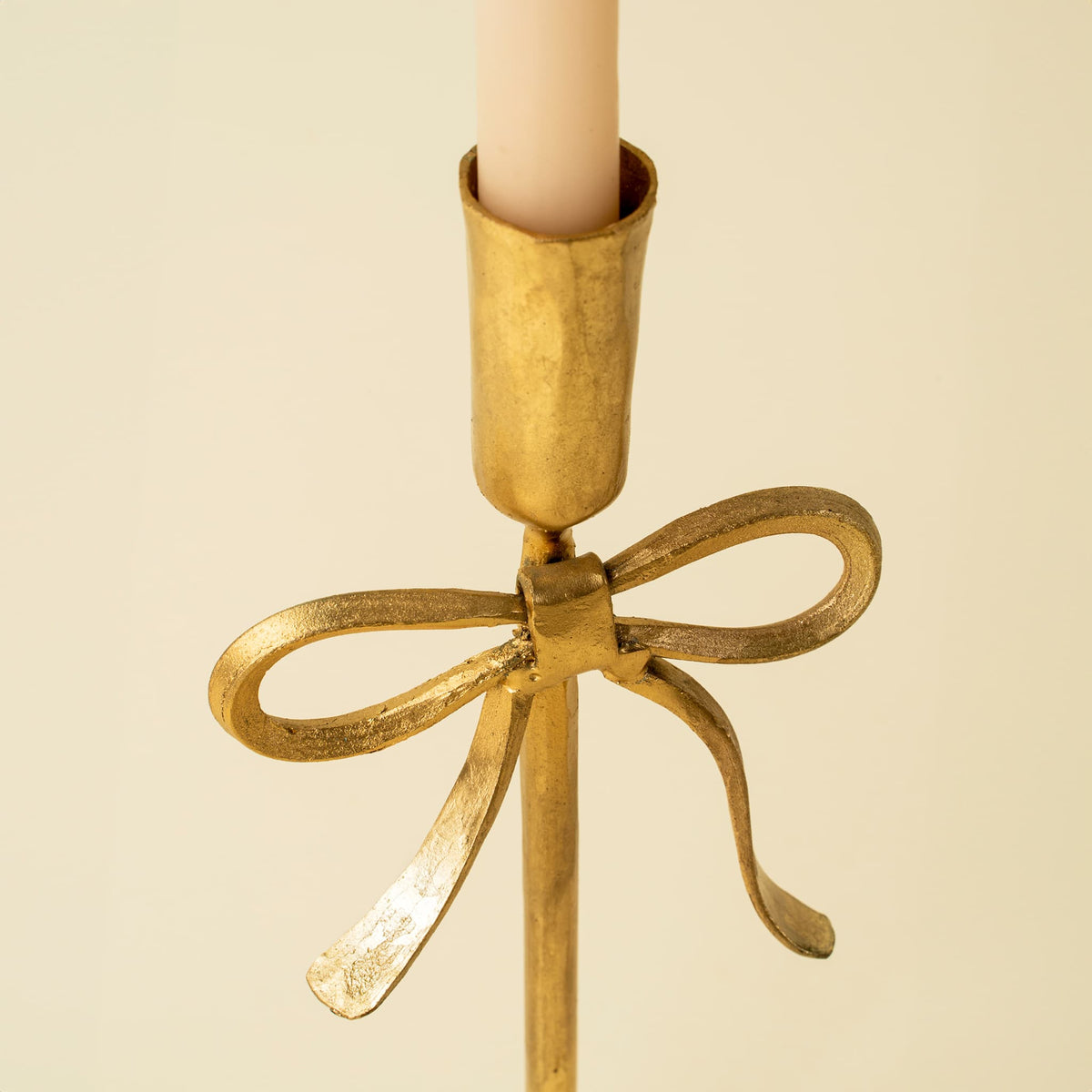 A close-up of a gold-toned metal candlestick holder with a decorative bow detail and a white candle inserted, set against a plain cream background.