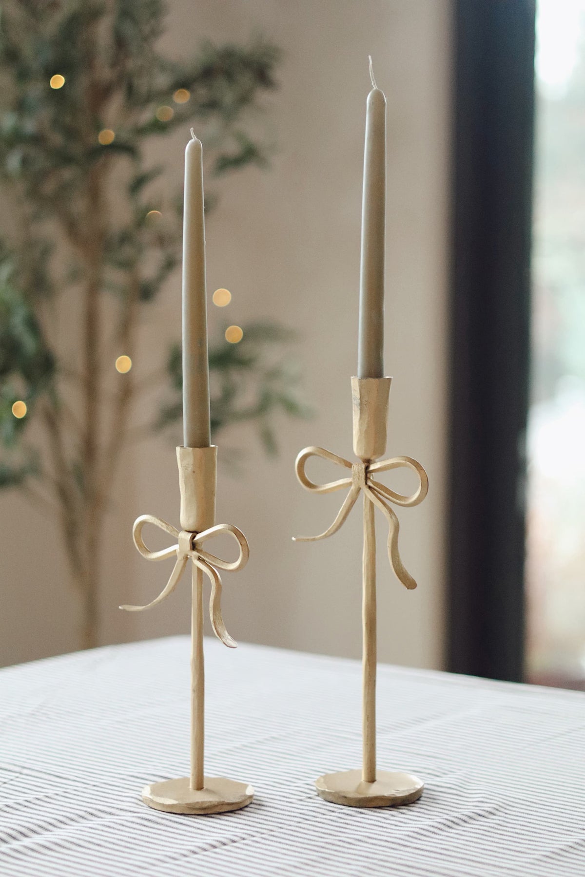 Two tall sage-coloured taper candles in matching holders with ribbon bow detail, displayed on a table in a softly lit indoor setting.