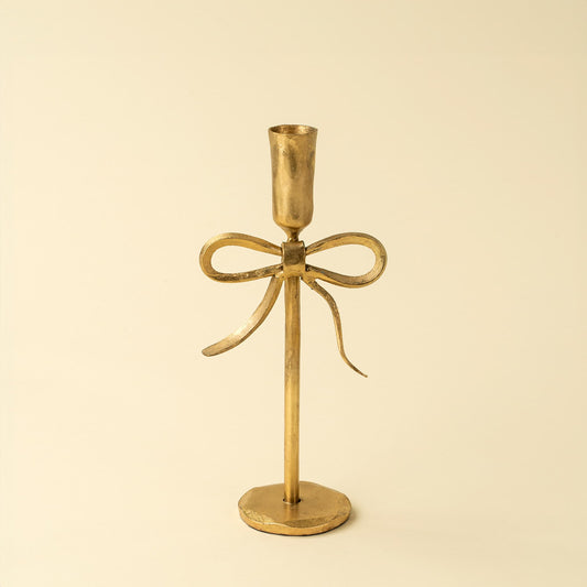 A single gold-toned metal candlestick holder, a vertical stem, a bow-shaped decorative element at the center, and a round base, photographed against a plain warm cream background.