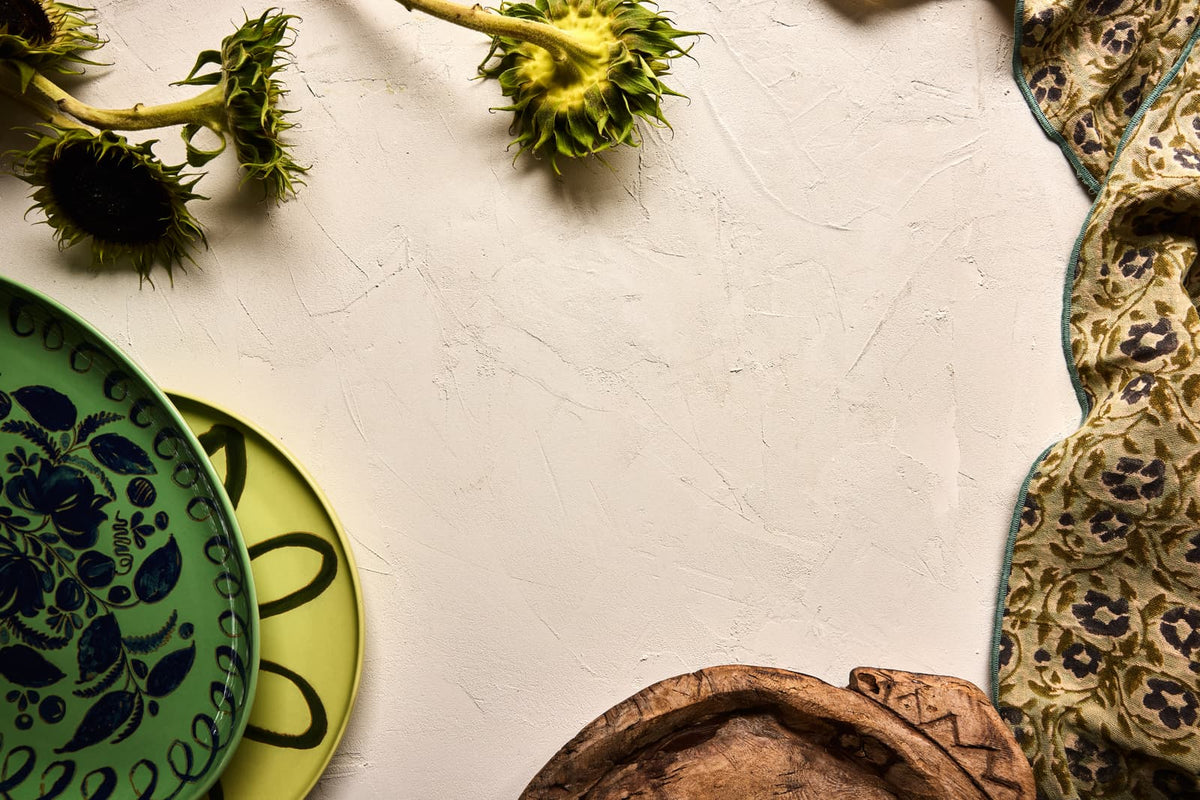 A styled flat-lay of decorative tableware on a textured off-white surface, featuring a green ceramic plate with a blue floral pattern, a yellow plate, a patterned beige fabric napkin or runner, and a brown carved wooden bowl, with sunflower heads used as props.