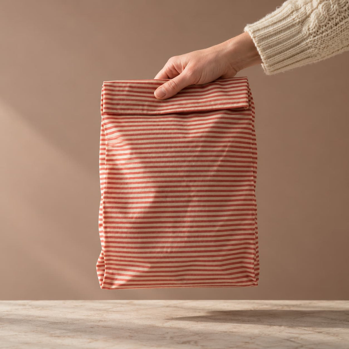 Hand holding red striped beeswax bread bag in front of neutral background with soft light.