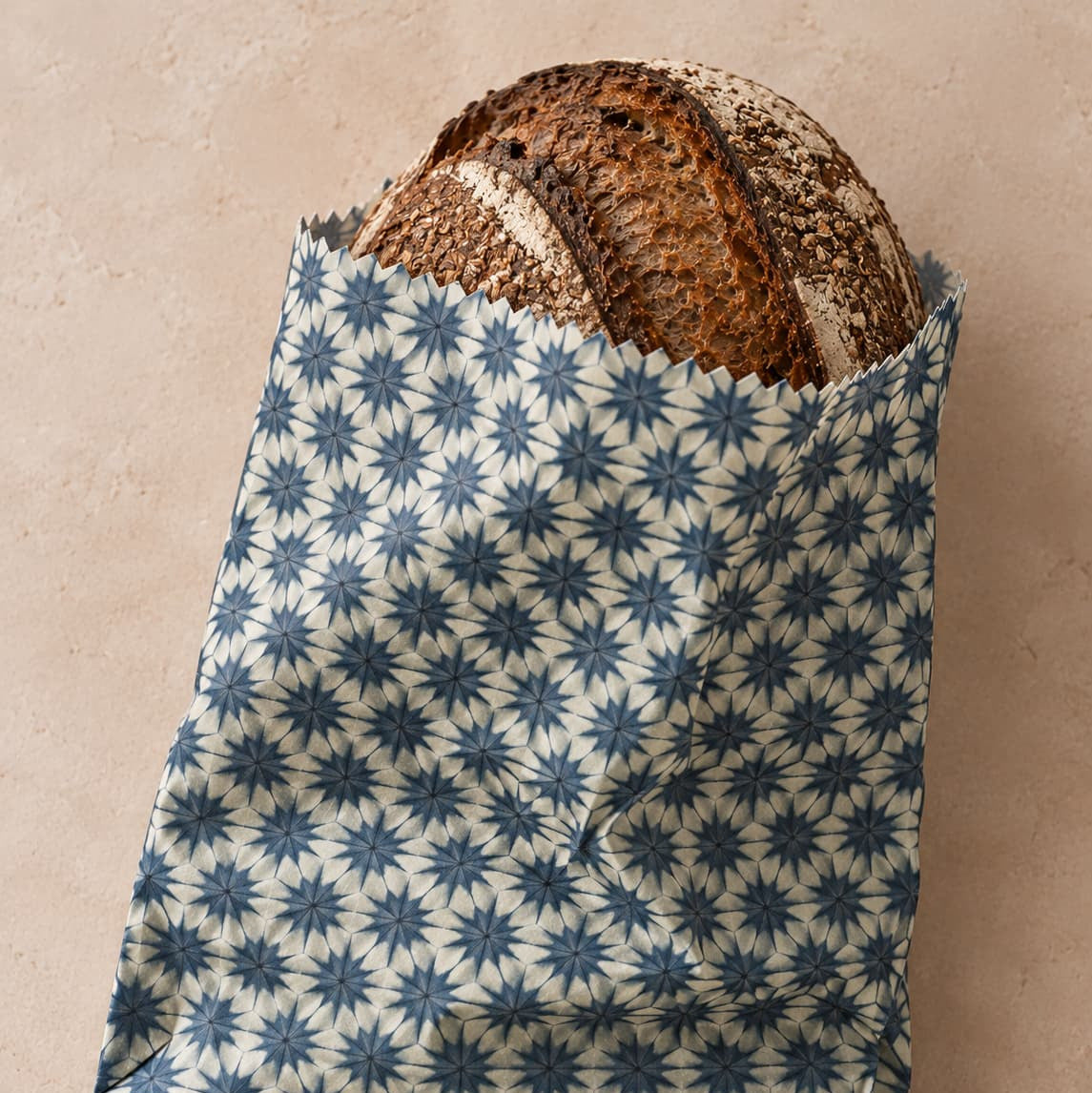 Sourdough loaf in a reusable beeswax bag in blue starburst fabric on a clean beige background.