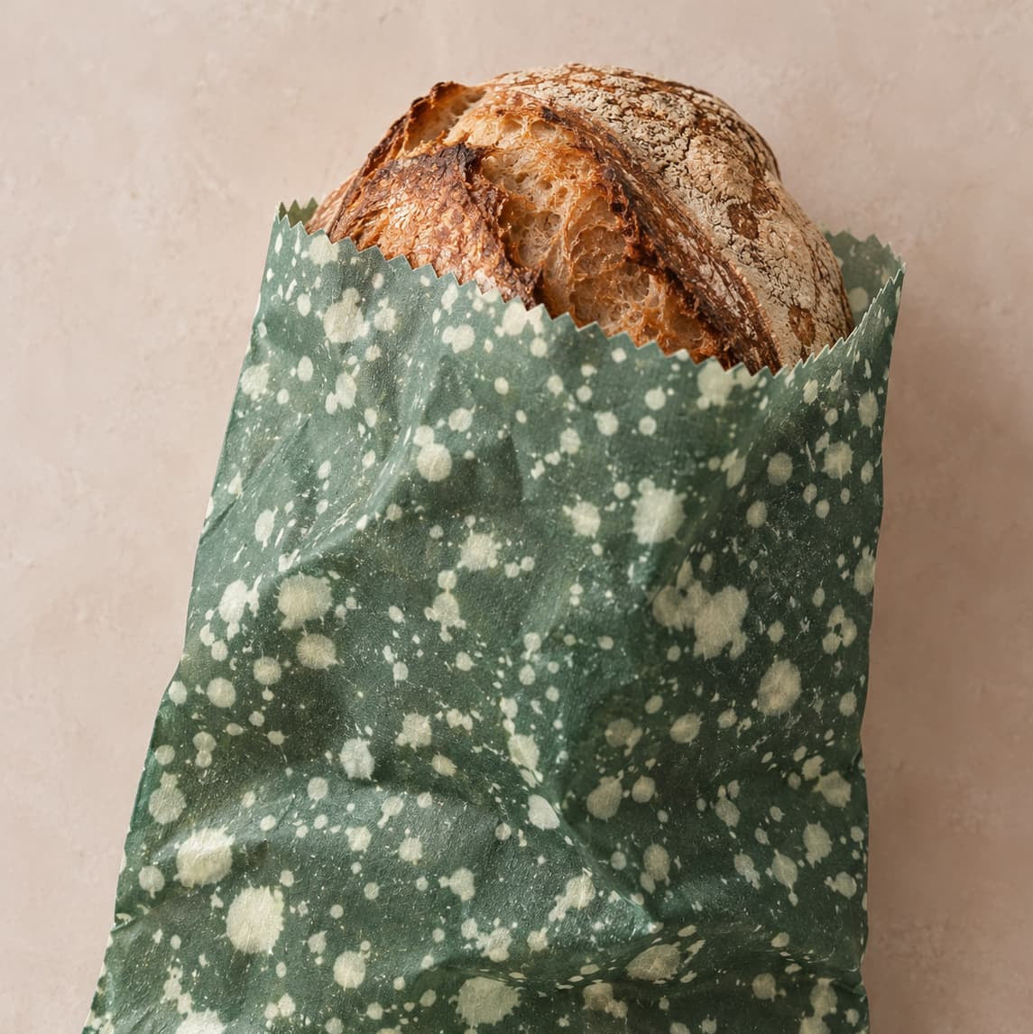Sourdough loaf in a reusable beeswax bag in dark green speckled fabric on a clean beige background.