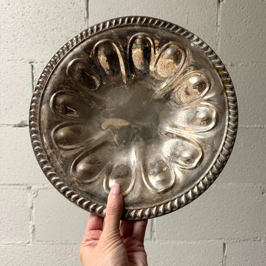 A round silver-toned metal plate with a decorative scalloped rim and raised petal-like indentations, held by a hand against a light gray painted brick wall.