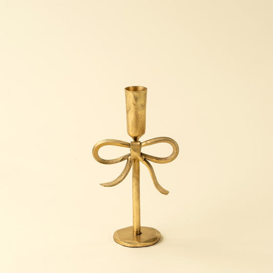 A small brass bow-shaped candlestick holder, set against a solid cream background.
