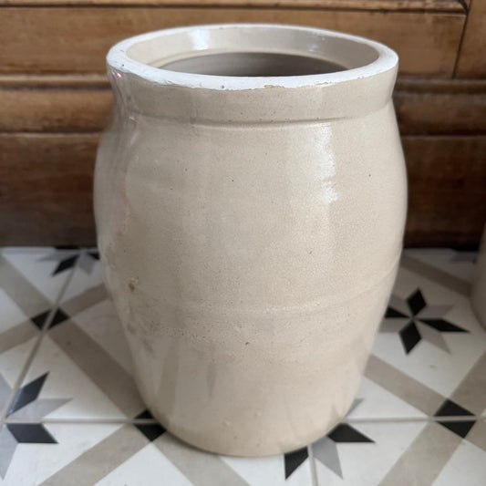 A stoneware crock in a warm beige glaze, standing on a black and white geometric tile floor against a rustic wooden door background.