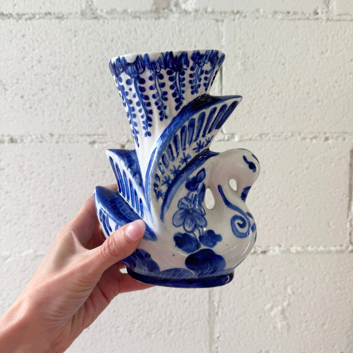 A hand holding a blue and white ceramic swan-shaped planter against a white painted brick wall.