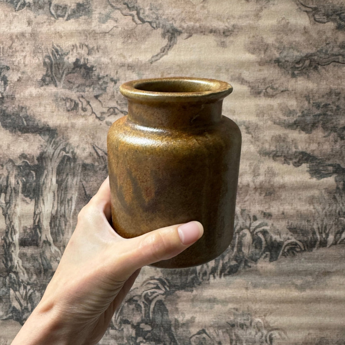 A hand holds a small brown ceramic jar with a wide flared rim in front of a textured, traditional-style landscape backdrop.