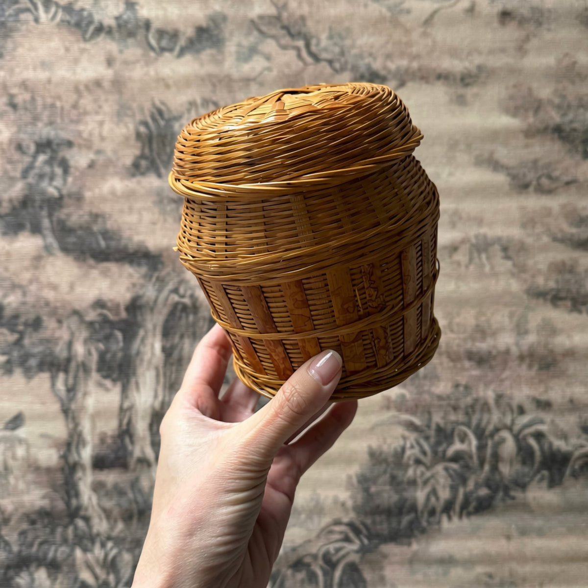 A hand holds a small woven rattan cylindrical basket with a fitted lid, set against a soft-focus traditional landscape backdrop.
