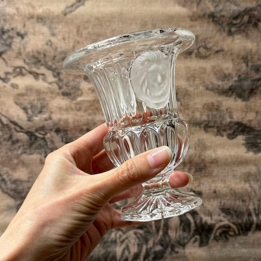 A hand holding a clear glass pedestal vase with a flared rim and a frosted lion motif on the front, set against a textured brown fabric background.