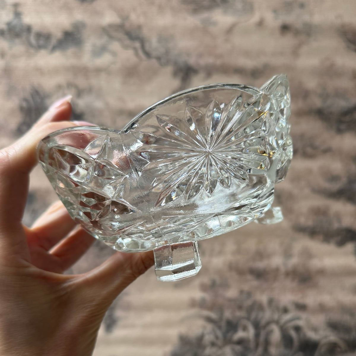 A hand holding a clear cut-glass bowl with a starburst pattern and small feet, set against a blurred brown patterned background.