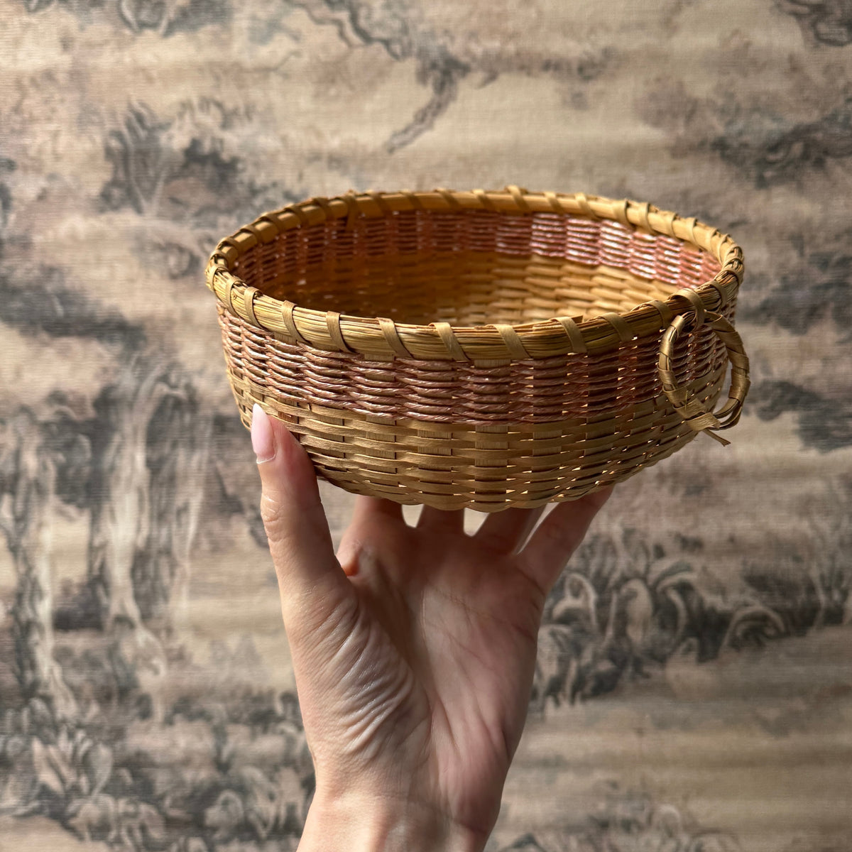 A hand holds a small round woven basket with a loop handle, shown against a large traditional landscape wall hanging.