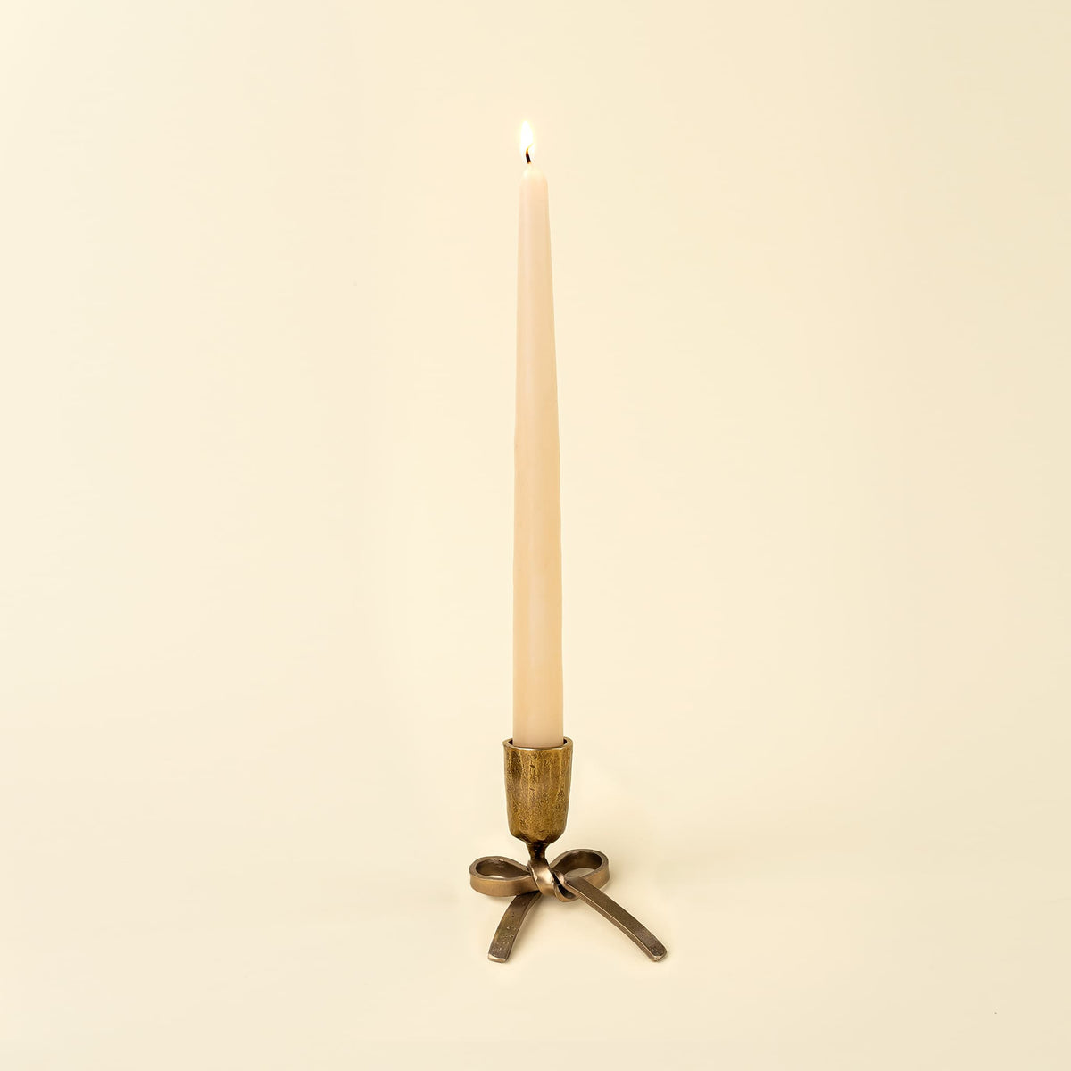 A single lit taper candle in a vintage-style metal holder with a bow-shaped base, photographed against a plain warm cream background.
