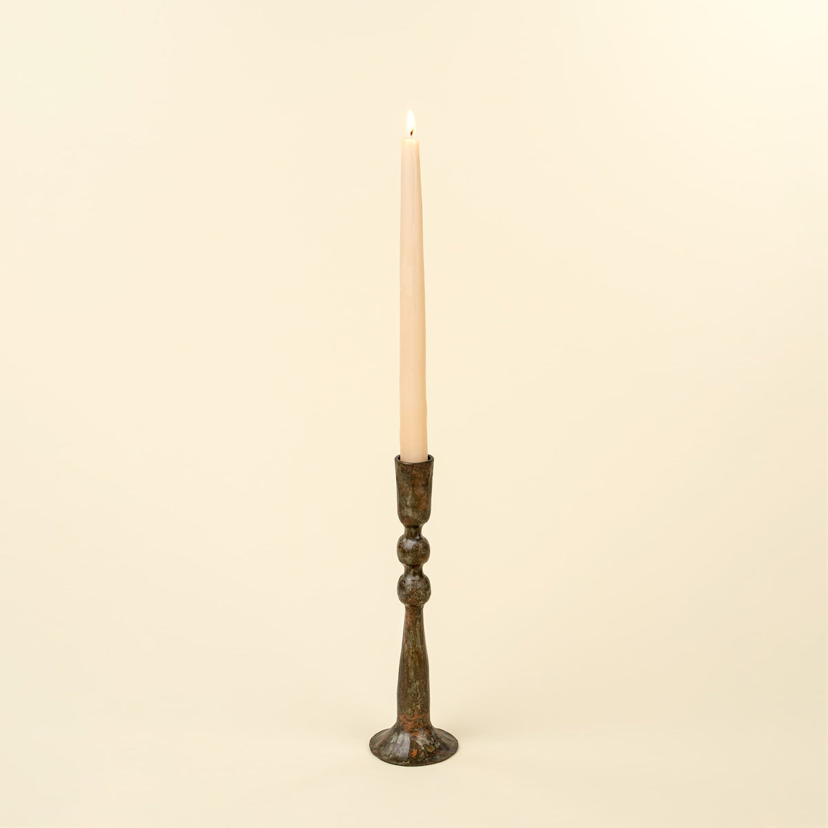 A single lit taper candle in a dark bronze metal candlestick holder, centered against a solid cream background.