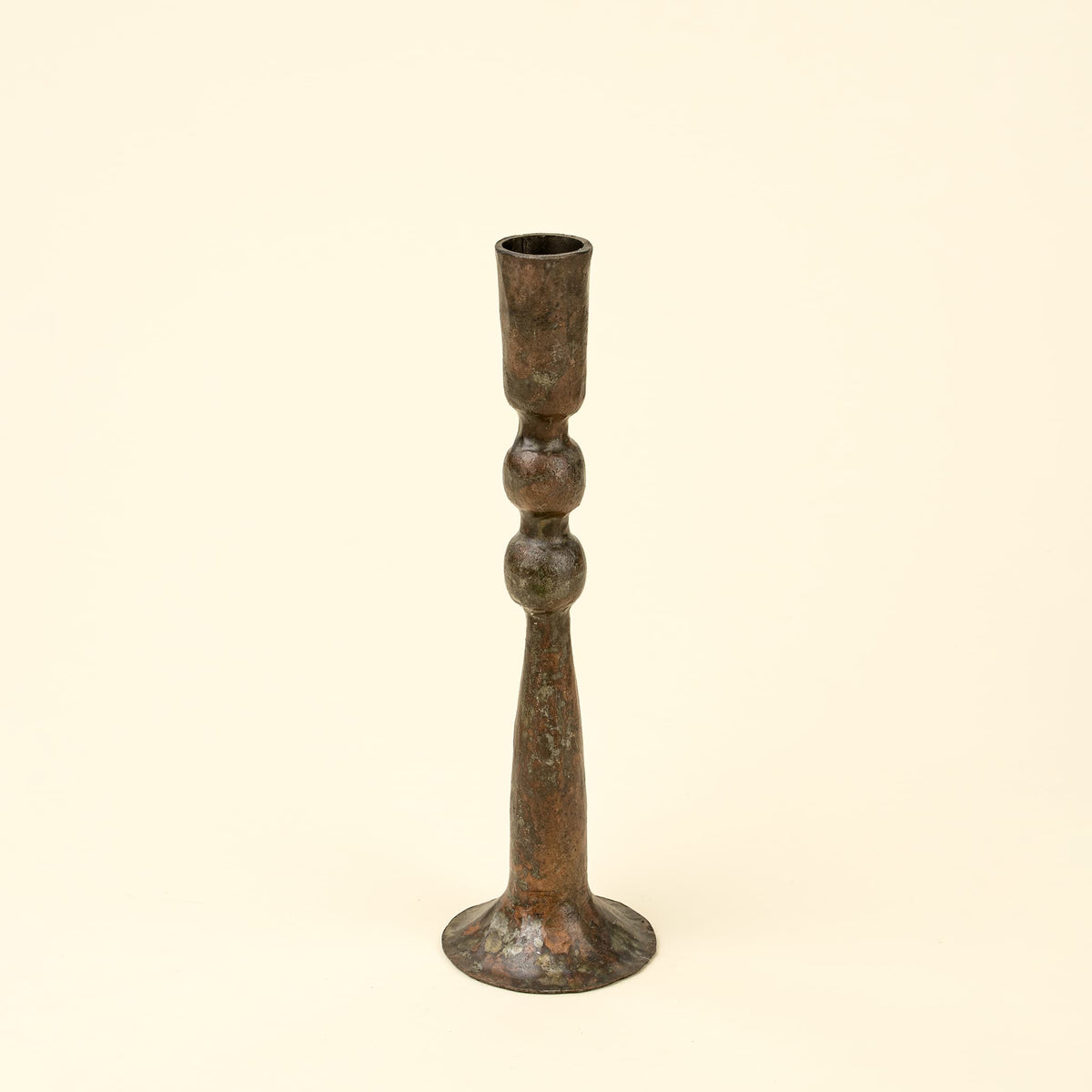A single rustic bronze candlestick holder with a textured, aged patina, centered against a solid cream-colored background.