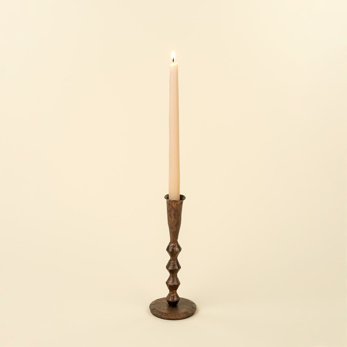 A single lit taper candle in a dark brown metal candlestick holder with a textured, ribbed design, set against a solid cream background.