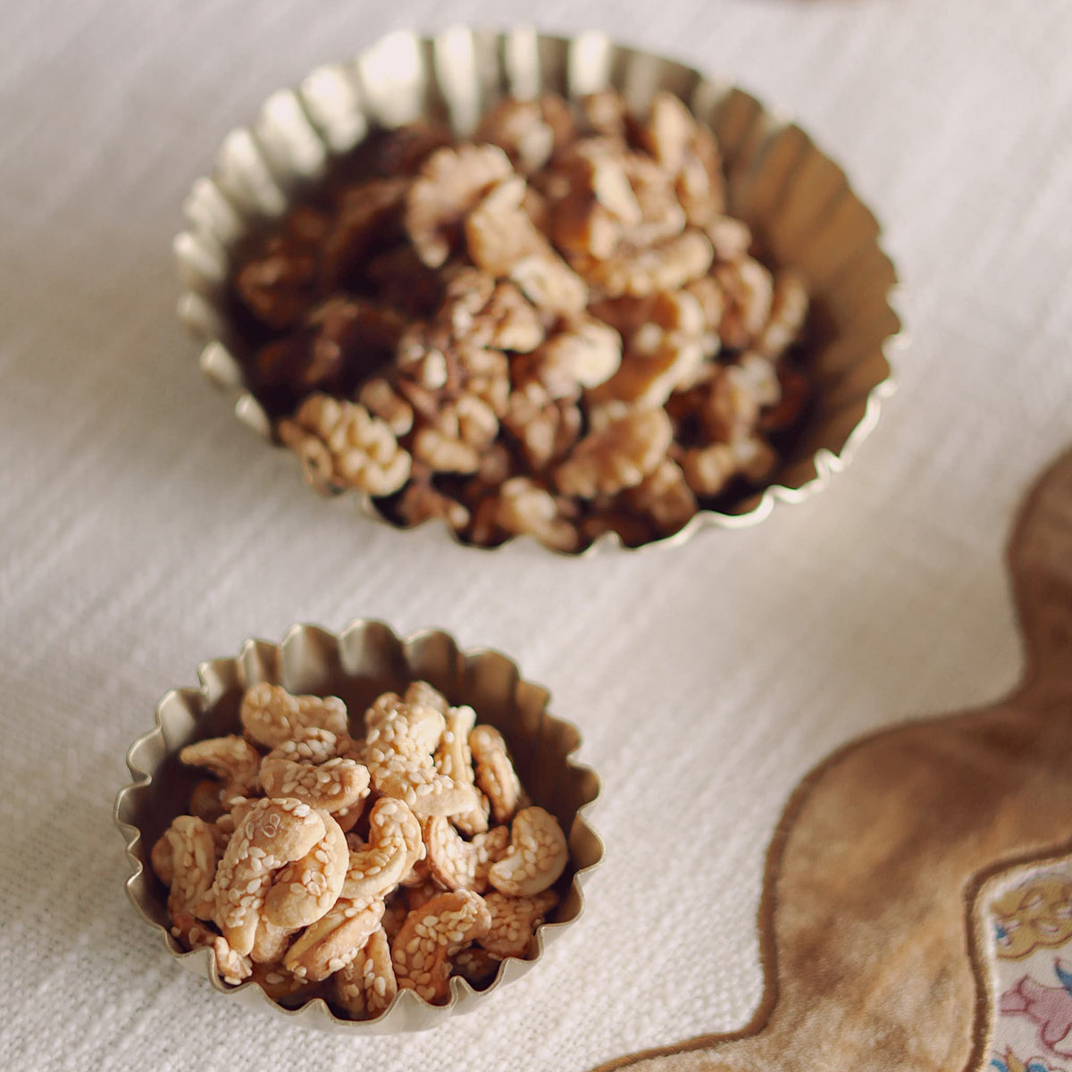 Two scalloped metallic bowls filled with candied nuts, set on a textured cream tablecloth next to a decorative brown paper cutout.