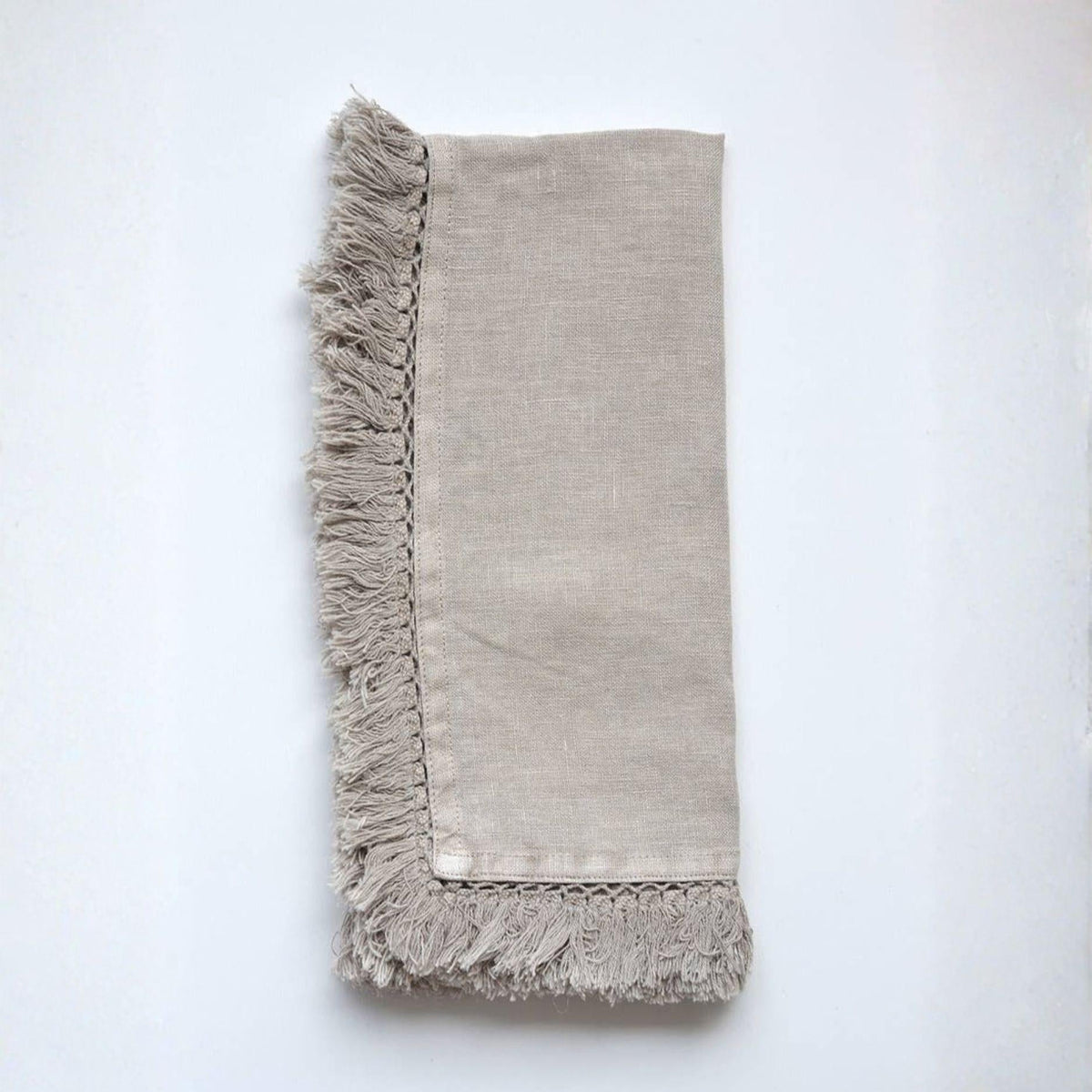 A folded light beige linen napkin with fringed edges, displayed flat against a clean white background.