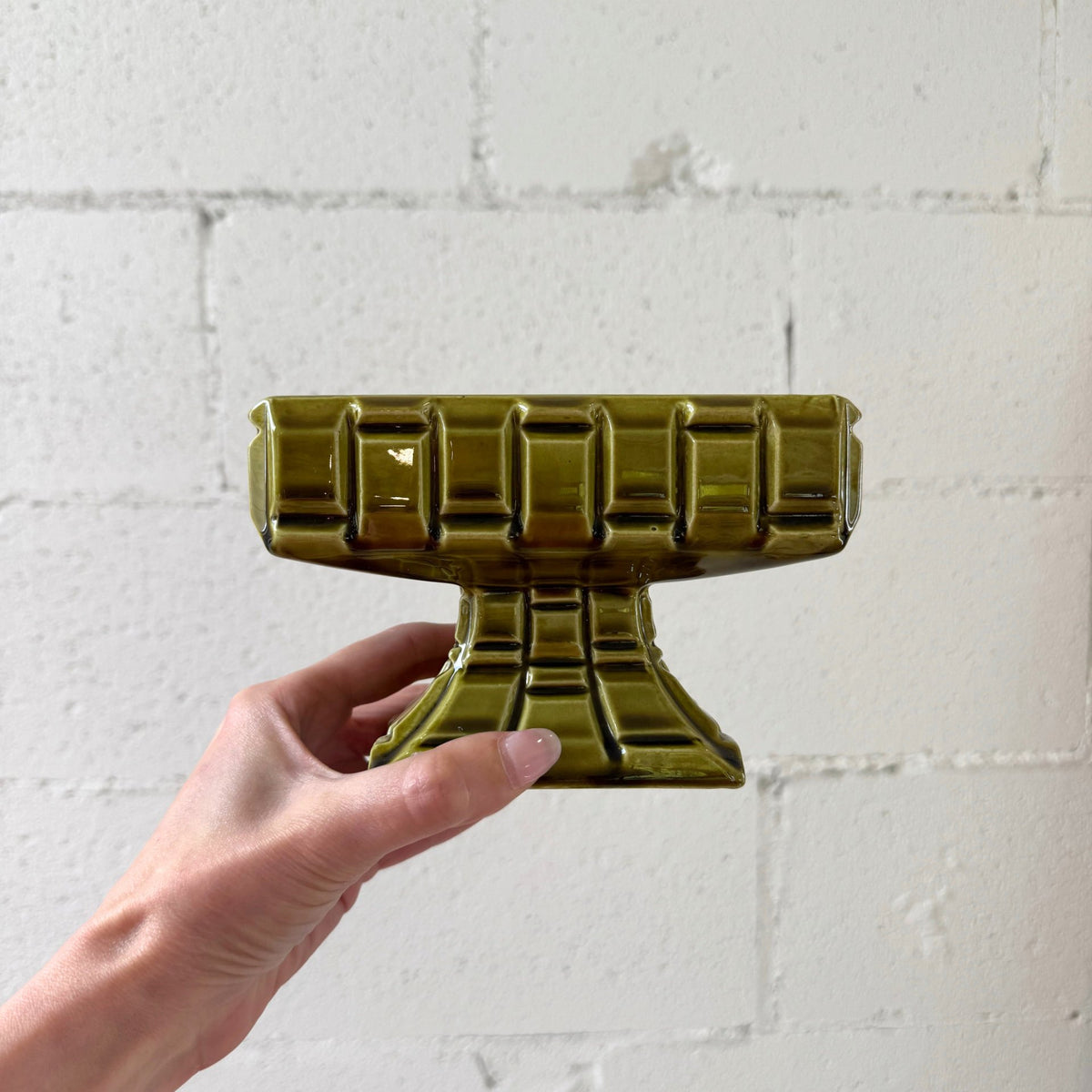 A hand holding a glossy olive green pedestal bowl with a ribbed, geometric design, set against a white painted brick wall.
