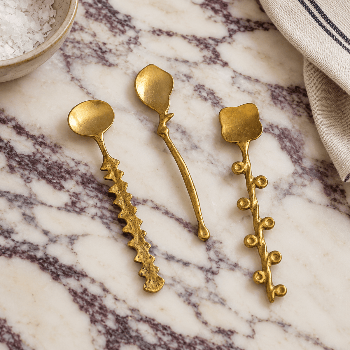Three decorative gold-toned metal spoons with organic-shaped handles are arranged on a calacatta viola marble surface.