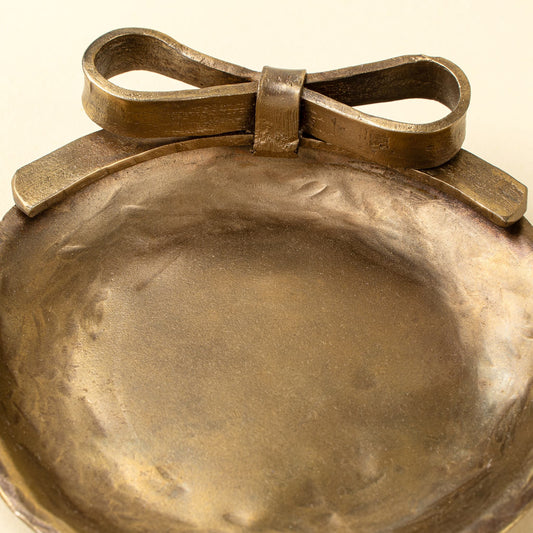 A close-up of a metallic aged brass bowl with a decorative bow detail along the rim, set against a plain light beige background.