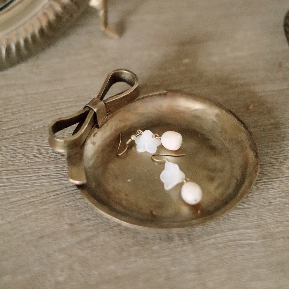 A small round metallic tray with a bow detail on the side, holding a pair of pearl earrings on a textured gray surface.