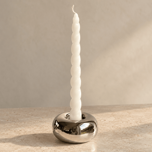 A white twisted taper candle standing upright in a polished silver metal holder on a light stone surface against a warm beige background.