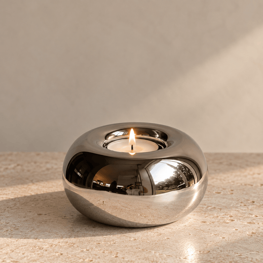 A polished chrome orb-shaped candle holder with a lit tealight candle inside, set on a textured beige stone surface against a neutral beige wall.