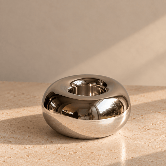 A polished chrome orb-shaped tealight holder, set on a beige stone surface against a matching beige wall with soft diagonal shadows.