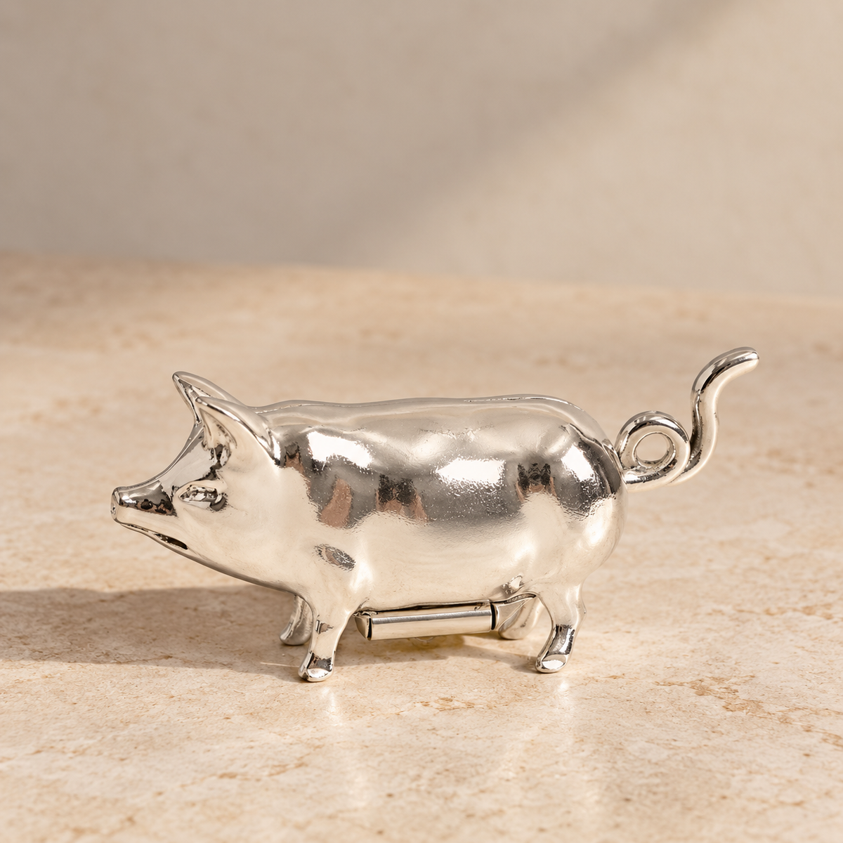 A polished silver metal pig-shaped object, likely a decorative figurine or small accessory, standing on a beige stone-like surface against a soft beige background.