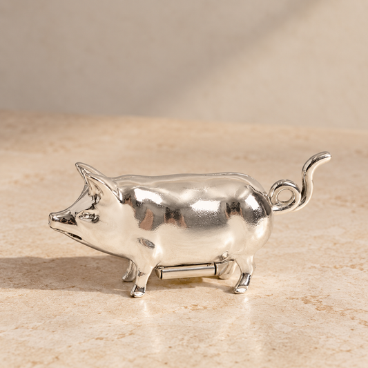 A polished silver metal pig-shaped object, likely a decorative figurine or small accessory, standing on a beige stone-like surface against a soft beige background.