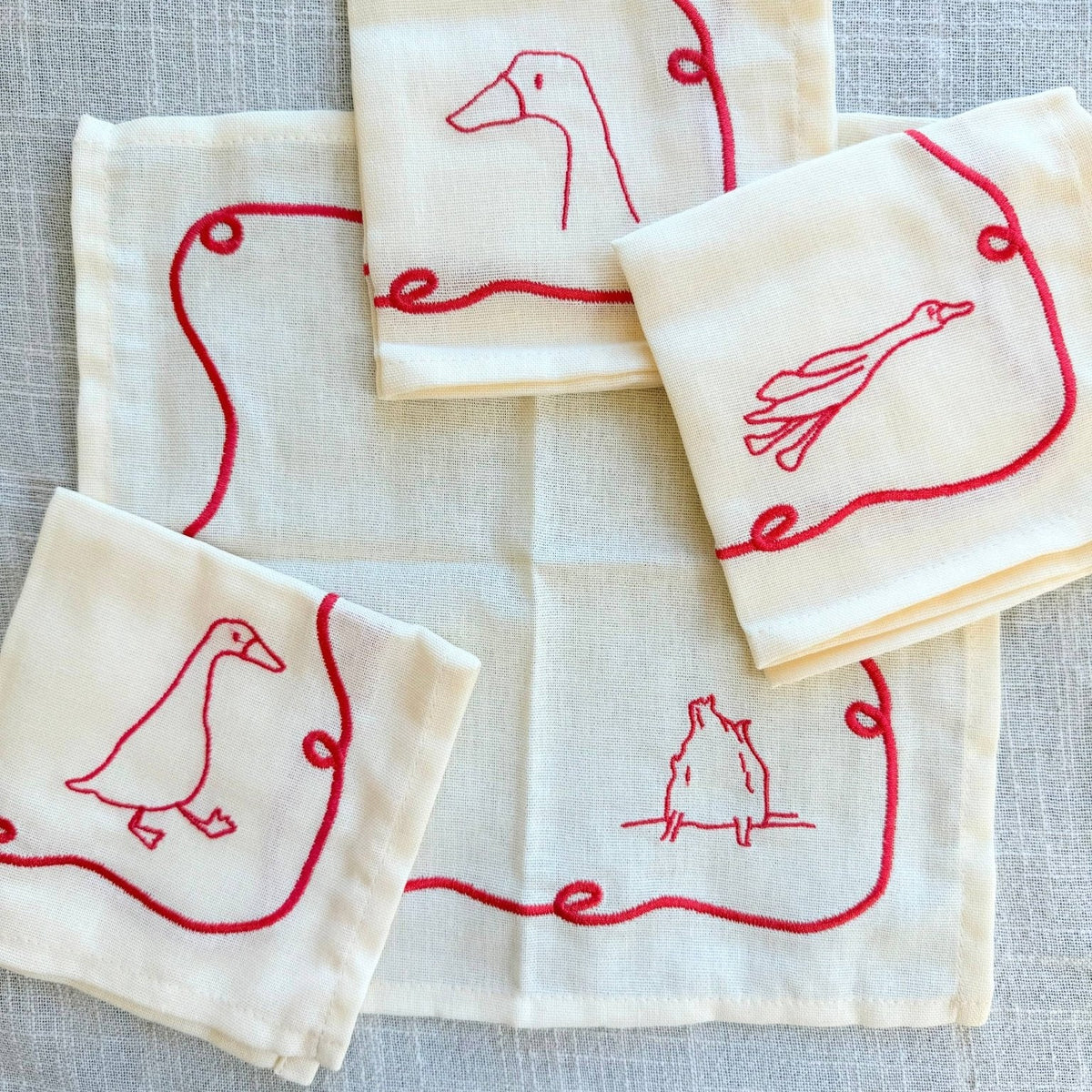 Four cream-colored linen napkins with red embroidered duck illustrations, arranged on a textured fabric background.
