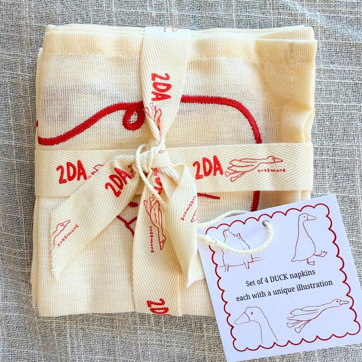 A folded set of four cream-coloured napkins tied with a matching ribbon embroidered with red ducks, displayed on a textured gray linen background.